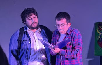 Hombre con barba y chaqueta de mezclilla escucha a otro con gafas y camisa de cuadros, en un ambiente iluminado.