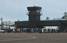 Grupo de personas en el aeropuerto, con vehículos estacionados, vestimenta variada, clima nublado y ambiente de actividad.