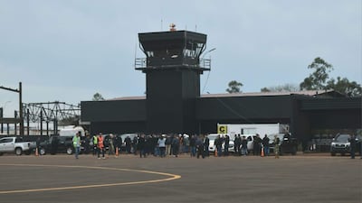 Grupo de personas en el aeropuerto, con vehículos estacionados, vestimenta variada, clima nublado y ambiente de actividad.