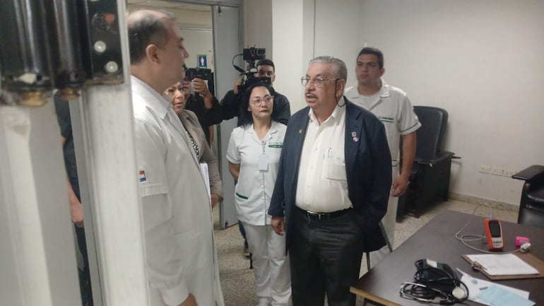 Hombre en bata blanca habla con otro en traje oscuro en hospital, mientras observan enfermeras y personal médico atentos.
