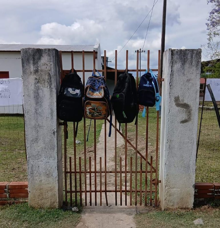 Cinco mochilas de distintos colores cuelgan de un portón de una institución educativa, en un ambiente vacío.