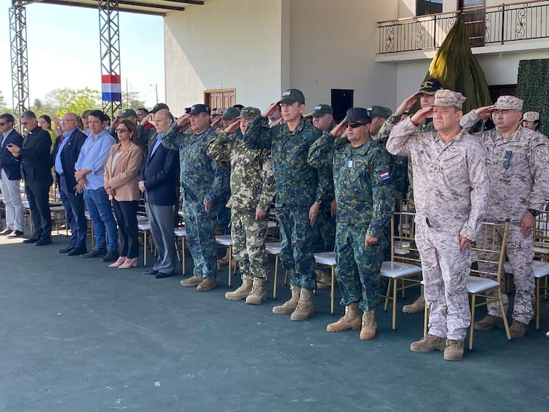 Grupo de adultos en uniforme militar y formal, con expresiones serias, en un ambiente ceremonial al aire libre.