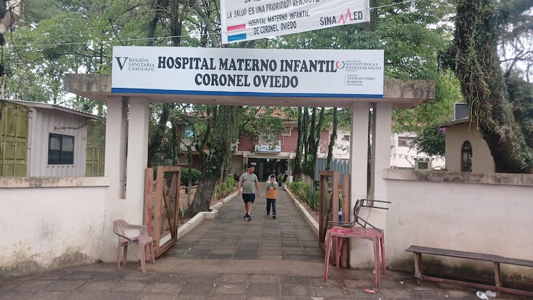 Adulto con camiseta gris y niño en sudadera naranja caminan hacia el hospital, rodeados de árboles y sillas de plástico vacías.