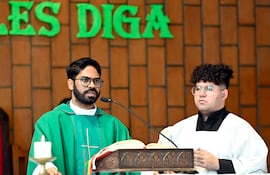 Sacerdote de vestimenta verde hablando con micrófono, asistido por un hombre joven con túnica blanca, en un entorno eclesiástico.