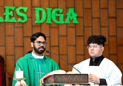 Sacerdote de vestimenta verde hablando con micrófono, asistido por un hombre joven con túnica blanca, en un entorno eclesiástico.