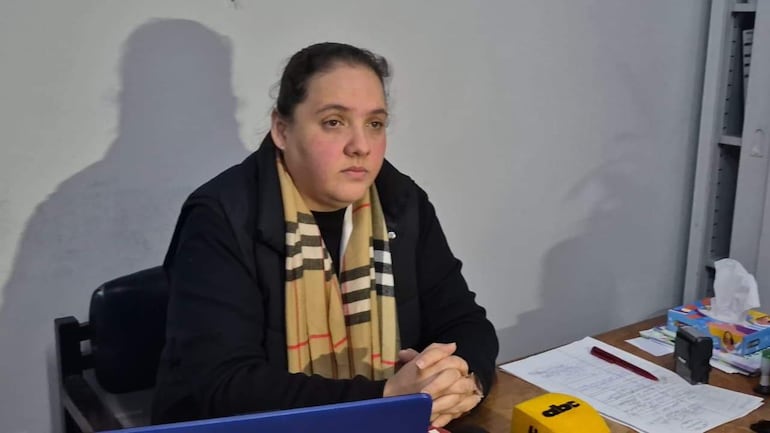 Mujer seria en chaqueta negra y bufanda a rayas, sentada en una mesa con computadora y papeles.