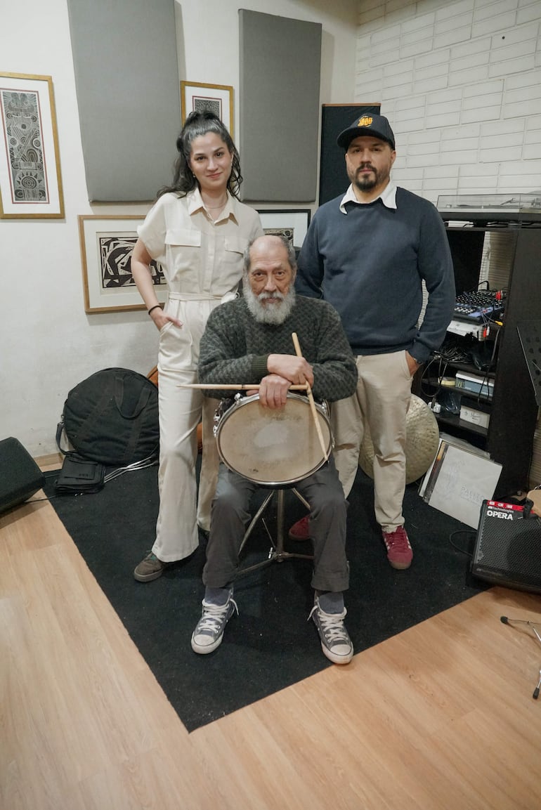Toti Morel, con suéter y baquetas, toca un tambor mientras Julieta y Víctor lo observan en un estudio musical colorido.