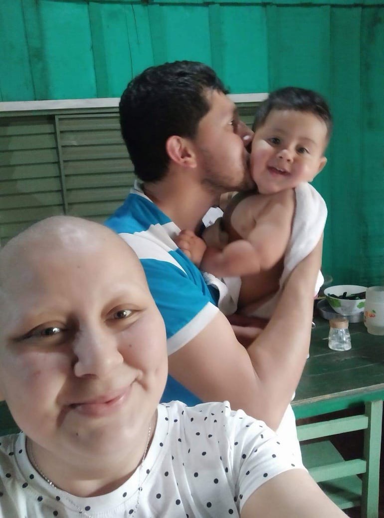 María Ojeda con camiseta blanca y cabello rapado sonríe, mientras un hombre con camiseta azul besa a un niño feliz en su hogar.