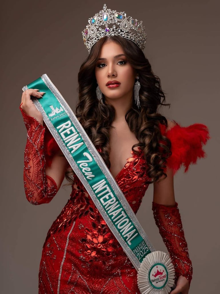 Katya Jara, con corona y vestido rojo brillante, sostiene banda que dice 'Reina Teen International' en un ambiente festivo.