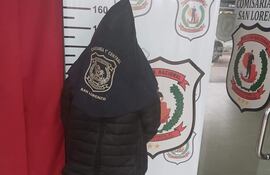 Persona vestida con chaqueta negra y capucha, de pie ante una pared con logo de la Policía Nacional en un ambiente interior.