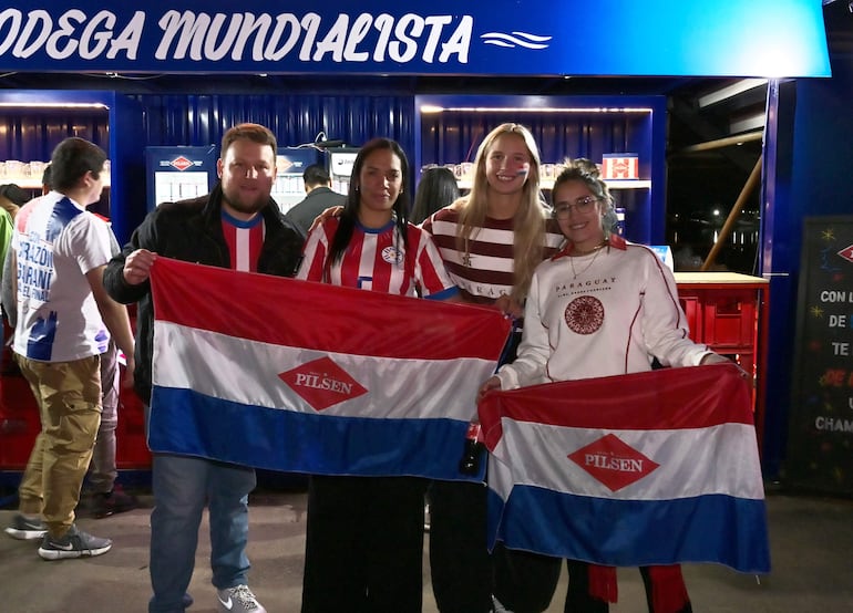 Cuatro personas sonrientes con banderas en un ambiente festivo, dos visten camisetas del Club Olimpia y una sostiene un refresco.