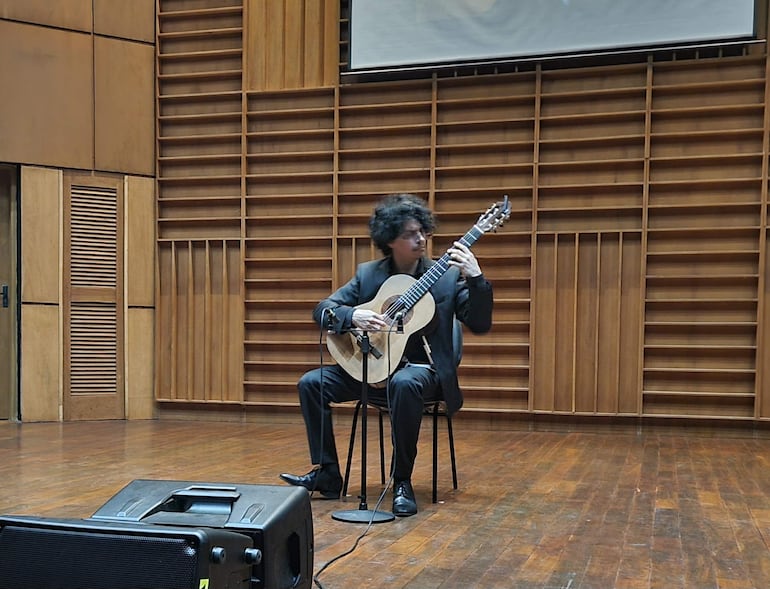 Alí Arango, vestido de traje oscuro, toca la guitarra con concentración en un escenario de madera durante su actuación.