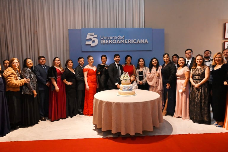 Veinticuatro personas en vestimenta formal rodean un pastel con el número 55 en un ambiente festivo y decorado con cortinas blancas.