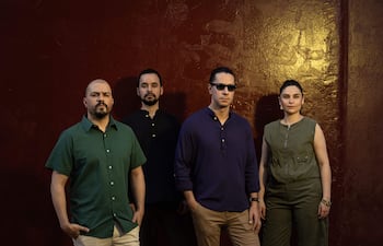 Cuatro personas sonríen frente a un fondo rojo; varón de camisa verde, otro en camiseta negra, hombre con camisa morada y mujer en overol verde.