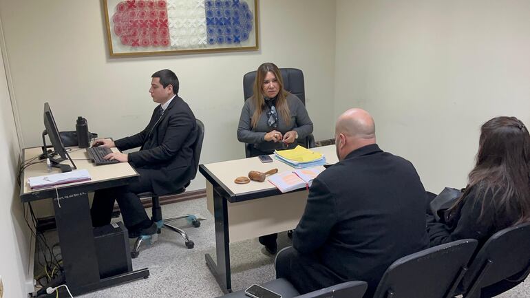 La jueza Clara Ruiz Díaz en el centro de la fotografía, mirando de frente. De espaldas el abogado Álvaro Arias, acompañado de una mujer. El expresidente del Indert, Ignacio Ortigoza, participando de una audiencia de imposición de medidas a través de un celular que está a lado del dactilógrafo del Poder Judicial.