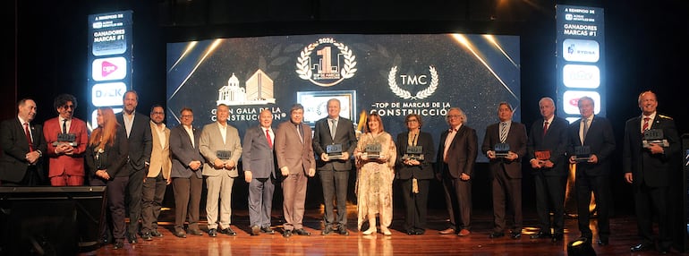 Grupo de premiados en gala, todos vestidos formalmente, algunos sosteniendo trofeos, en un escenario elegante.