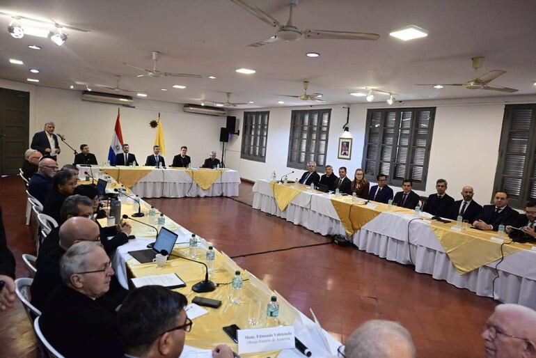 Grupo de personas en traje formal y eclesiástico, escuchando atentamente a un orador de pie en un ambiente formal.