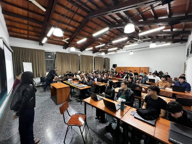 Dos jóvenes interactúan en un aula con techos altos; estudiantes atentos con computadoras. Ambiente académico y de debate.