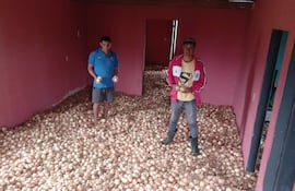 Dos hombres sostienen cebollas en sus manos dentro de una habitación con piso cubierto de cebollas. Uno viste camiseta azul y shorts, el otro, chaqueta roja y camiseta amarilla.