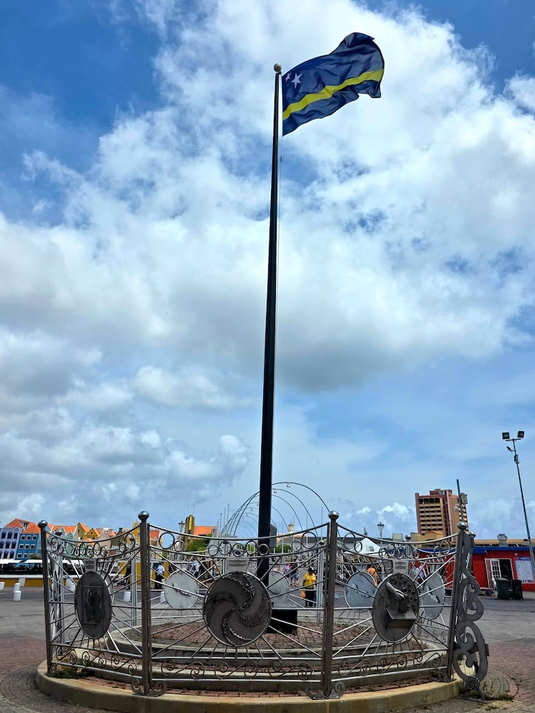 Bandera azul y amarilla ondea en un mástil en un parque de Willemstad, con edificios coloridos de fondo y cielo nublado.