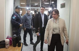 Hombre en blazer negro junto a mujer de atuendo claro, observados por un hombre con camisa de mezclilla y un oficial de policía.