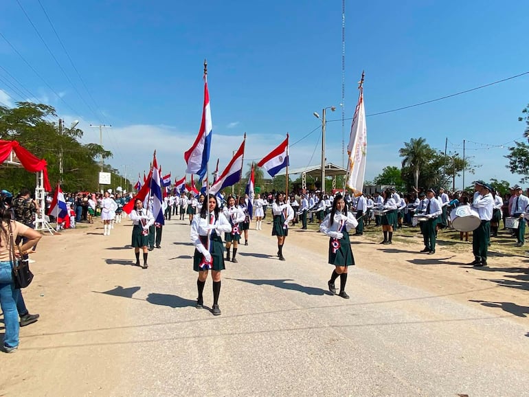 Estudiantes en uniforme blanco y pantalones oscuros marchan mientras sostienen banderas paraguayas en un ambiente festivo.