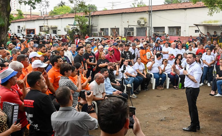 Luis Bello habla con micrófono, vestido de blanco, rodeado de público que usa camisetas de colores naranja y negro, escuchando atentamente.
