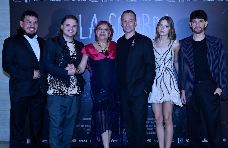 Seis personas sonrientes en evento de cine; un hombre de traje negro y mujeres con vestidos coloridos en fondo decorado.