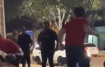 Hombre con camiseta roja en primer plano, rodeado por otros hombres con ropa oscura, en un ambiente urbano.