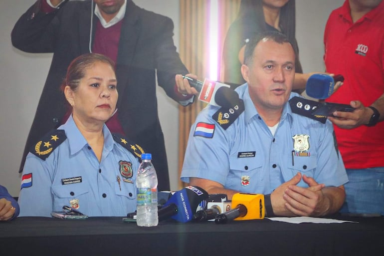 Diosnel Alarcón y mujer con uniformes policiales, frente a mesa con micrófonos y botellas de agua, en ambiente formal.