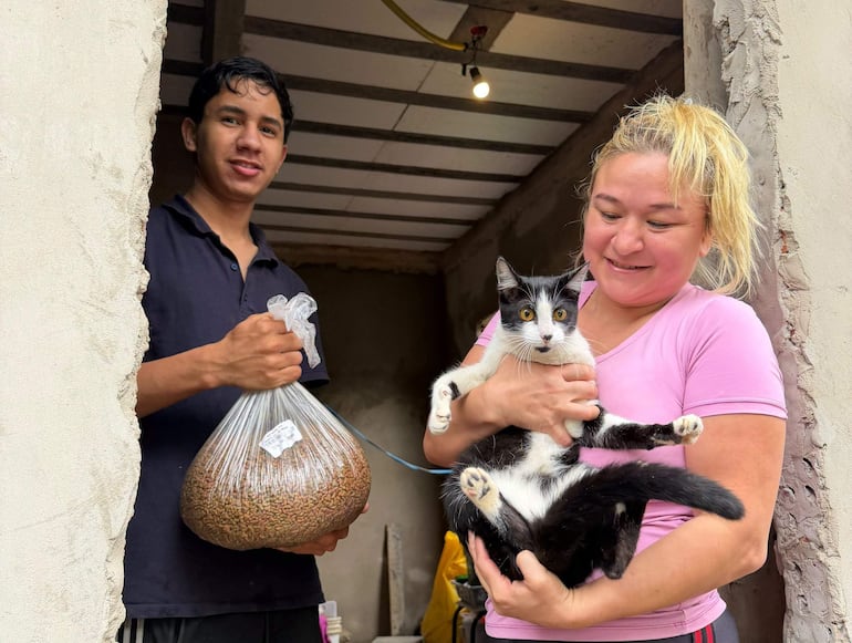 Joven con camiseta oscura sostiene bolsa de alimento, mientras mujer sonriente con coleta abraza un gato negro y blanco.