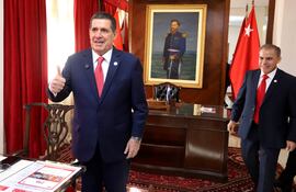 Horacio Cartes sonriente con traje azul y corbata roja, levantando el pulgar en una oficina formal con banderas y cortinas rojas.