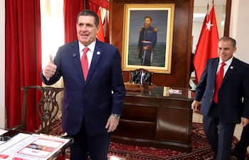 Horacio Cartes sonriente con traje azul y corbata roja, levantando el pulgar en una oficina formal con banderas y cortinas rojas.