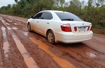 Automóvil blanco con barro aparcado en camino en mal estado, rodeado de vegetación densa.