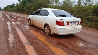 Automóvil blanco con barro aparcado en camino en mal estado, rodeado de vegetación densa.