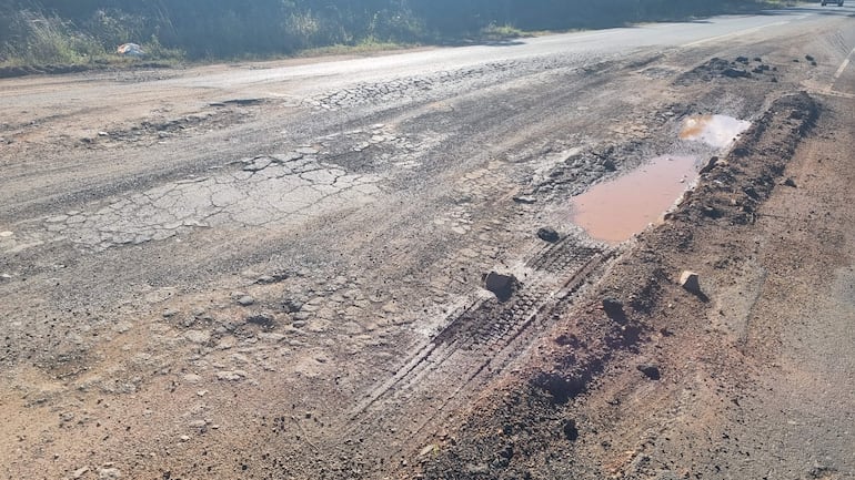 Carretera en mal estado con baches y charcos, rodeada de vegetación, sin vehículos ni personas visibles.