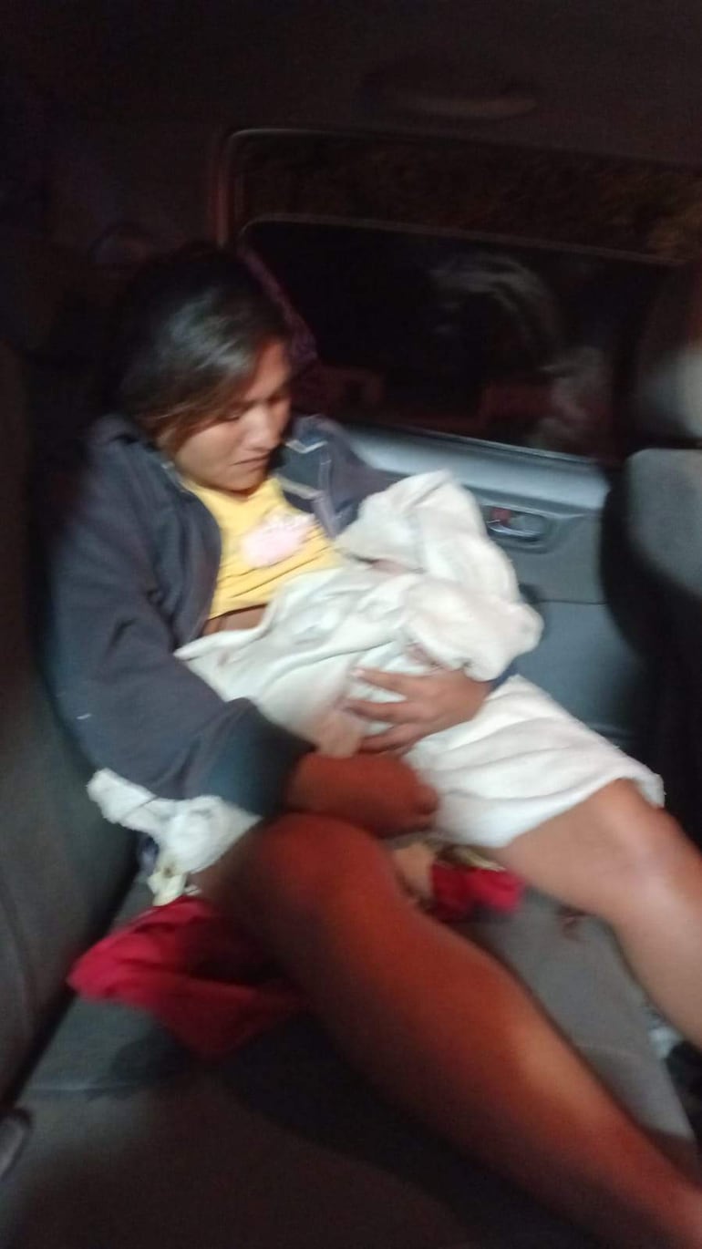 Mujer indígena con cabello oscuro y camiseta amarilla sostiene a su bebé envuelto en una manta blanca en un vehículo oscuro.