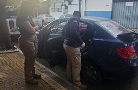 Un funcionario de seguridad inspecciona un automóvil azul dañado, mientras otro observa con los brazos cruzados en una calle.
