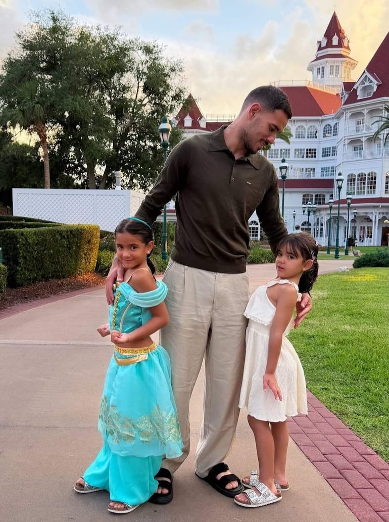 Antonio Sanabria, con camiseta oscura y pantalones claros, sostiene a una niña de disfraz azul, mientras la otra niña, en vestido blanco, sonríe curiosa.