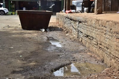 Contenedor marrón de basura en primer plano, área con agua estancada y barro, pared de ladrillo expuesto al fondo.