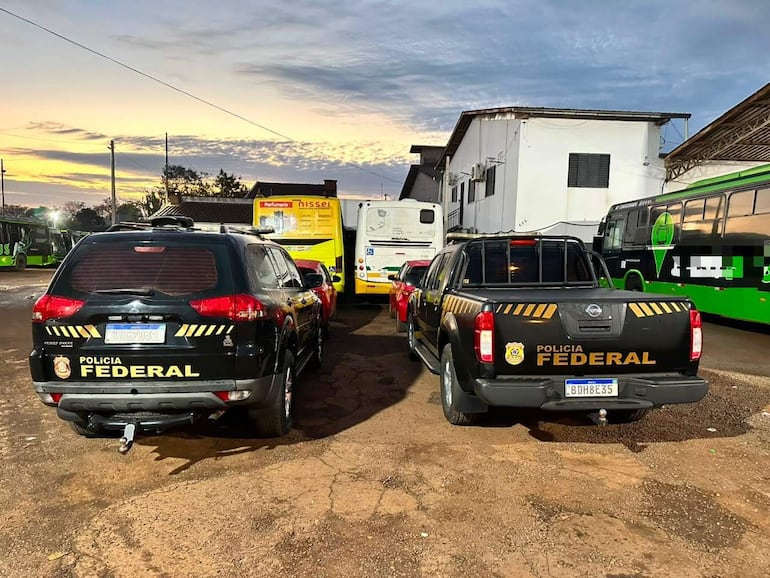 Vehículos de la Policía Federal, uno SUV y otro pick-up, en una escena tranquila al atardecer, rodeados de autobuses y un edificio en el fondo.