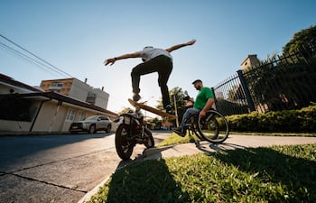 Hombre en silla de ruedas con camiseta verde observa a skater en acción en un entorno urbano soleado.