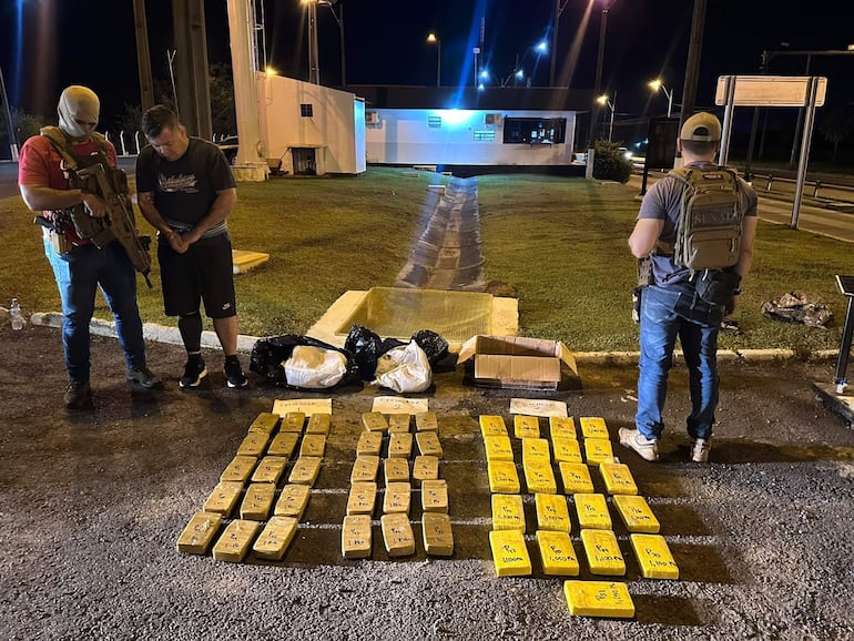 Hombre detenido con manos atadas, custodiado por agente armado con chaleco táctico, rodeado de paquetes de droga en control nocturno.