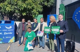 Seis personas en evento al aire libre, sosteniendo carteles sobre reciclaje junto a un contenedor de reciclaje.