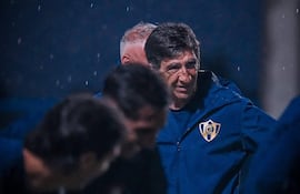 Hombre en posición de entrenador dirige práctica de fútbol con indumentaria oscura bajo la lluvia en un campo iluminado.