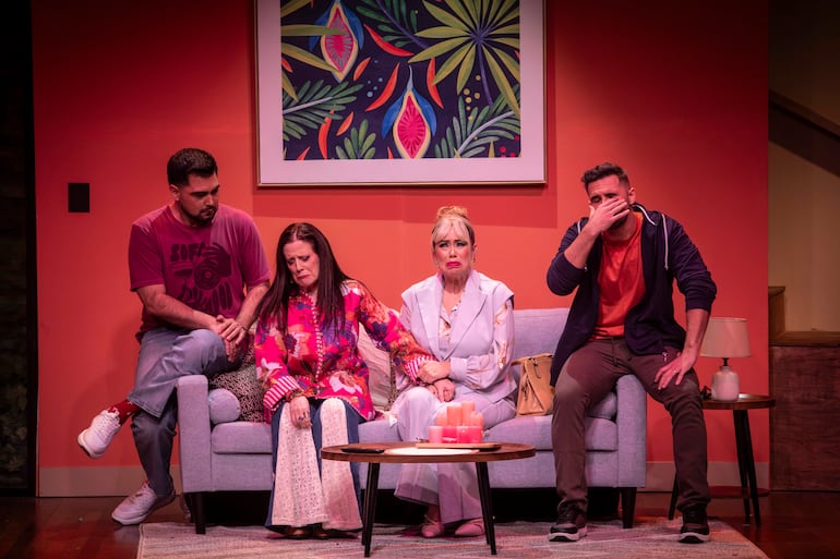 Mujer vestida de blanco con expresión de tristeza, acompañada de tres personas en un sofá, en escenario teatral con paredes naranjas.