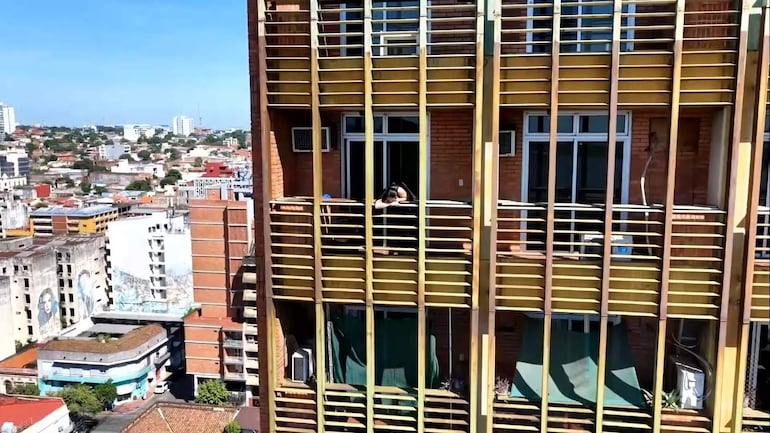 Persona femenina en camiseta negra relajándose en el balcón del edificio Ahorros Paraguayos, cielo despejado de fondo.