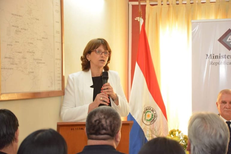 Gilda Villalba Tottil en chaqueta blanca habla con micrófono, rodeada de espectadores en ambiente institucional con bandera paraguaya.
