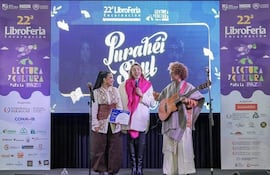 Tres personas en el escenario de la Libroferia: mujer con cabello recogido, mujer con bufanda rosa y hombre con guitarra, todos sonriendo juntos.