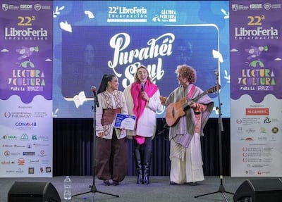 Tres personas en el escenario de la Libroferia: mujer con cabello recogido, mujer con bufanda rosa y hombre con guitarra, todos sonriendo juntos.